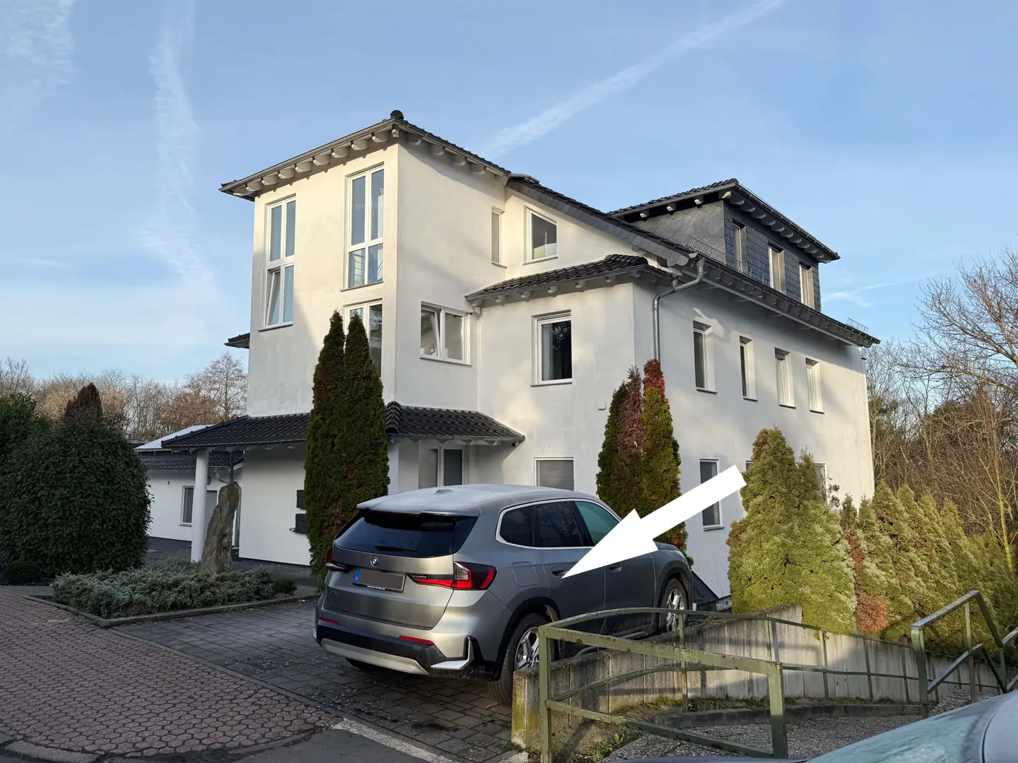 A modern, white, multi-story house with a gray BMW parked in front, indicated by a white arrow.