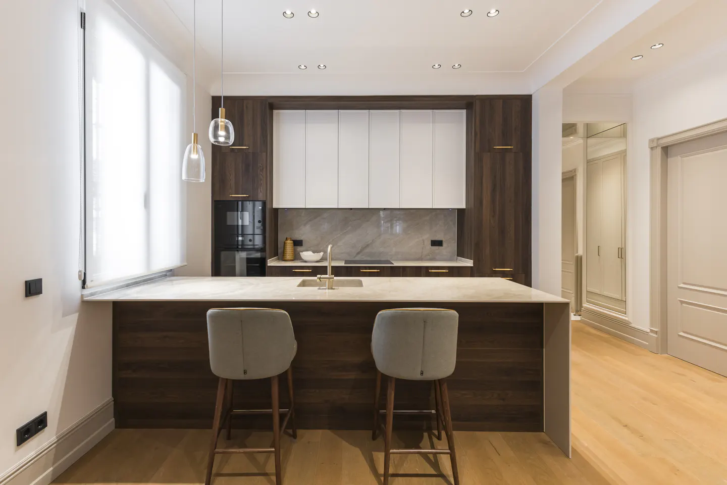 A modern kitchen with dark wood and white cabinets, a marble countertop island with two gray stools, and wood floors.