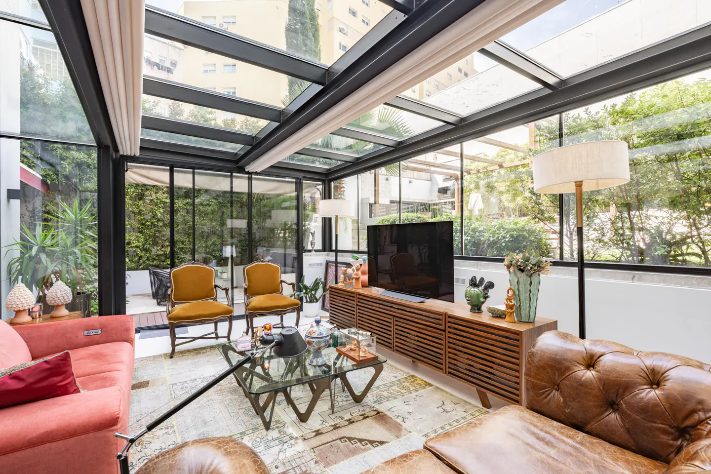 Bright sunroom with glass ceiling, brown leather sofa, coral couch, and mustard chairs. A TV sits on a wooden console.