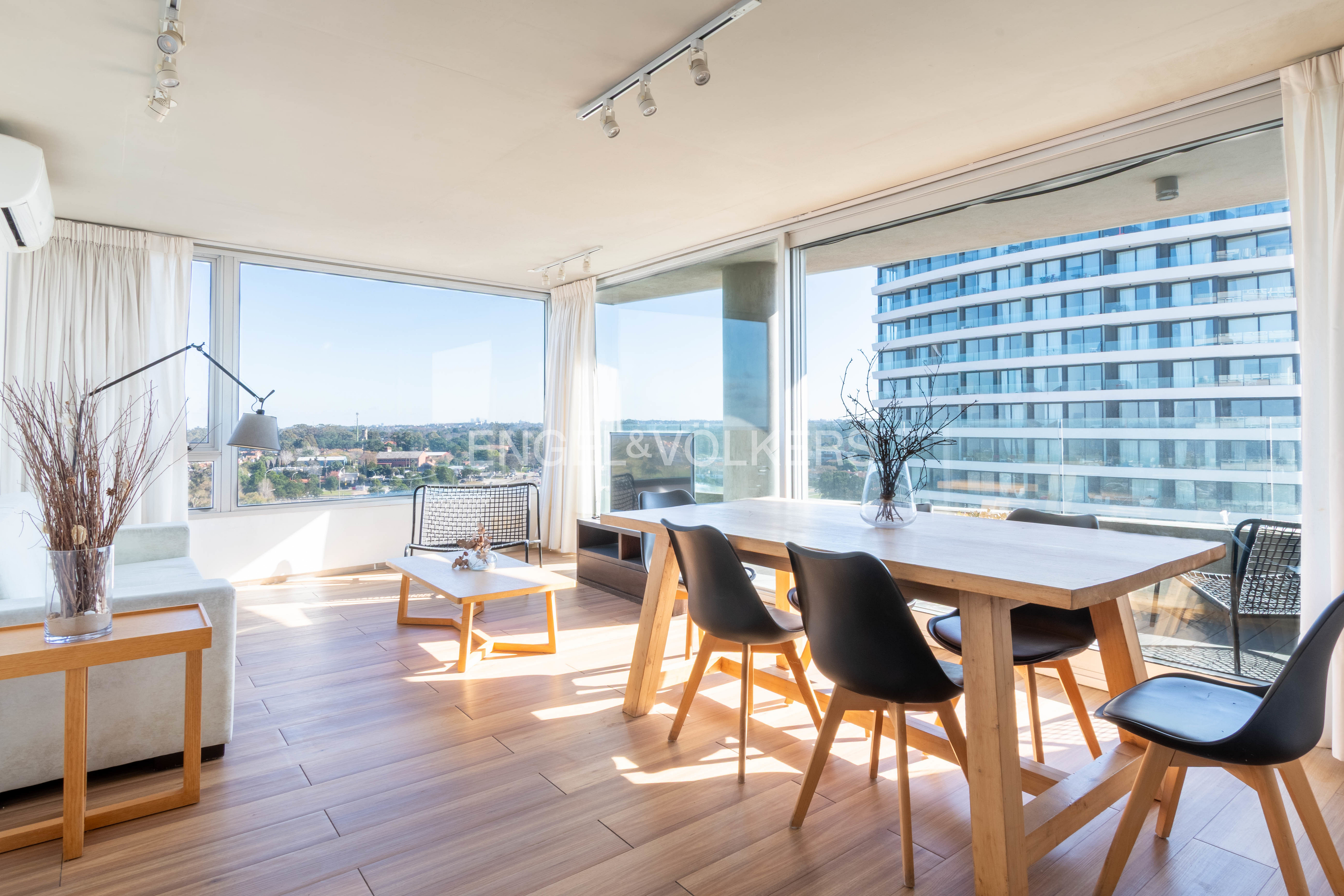 Bright living and dining area with wood floors, floor-to-ceiling windows, modern furniture, and city views.