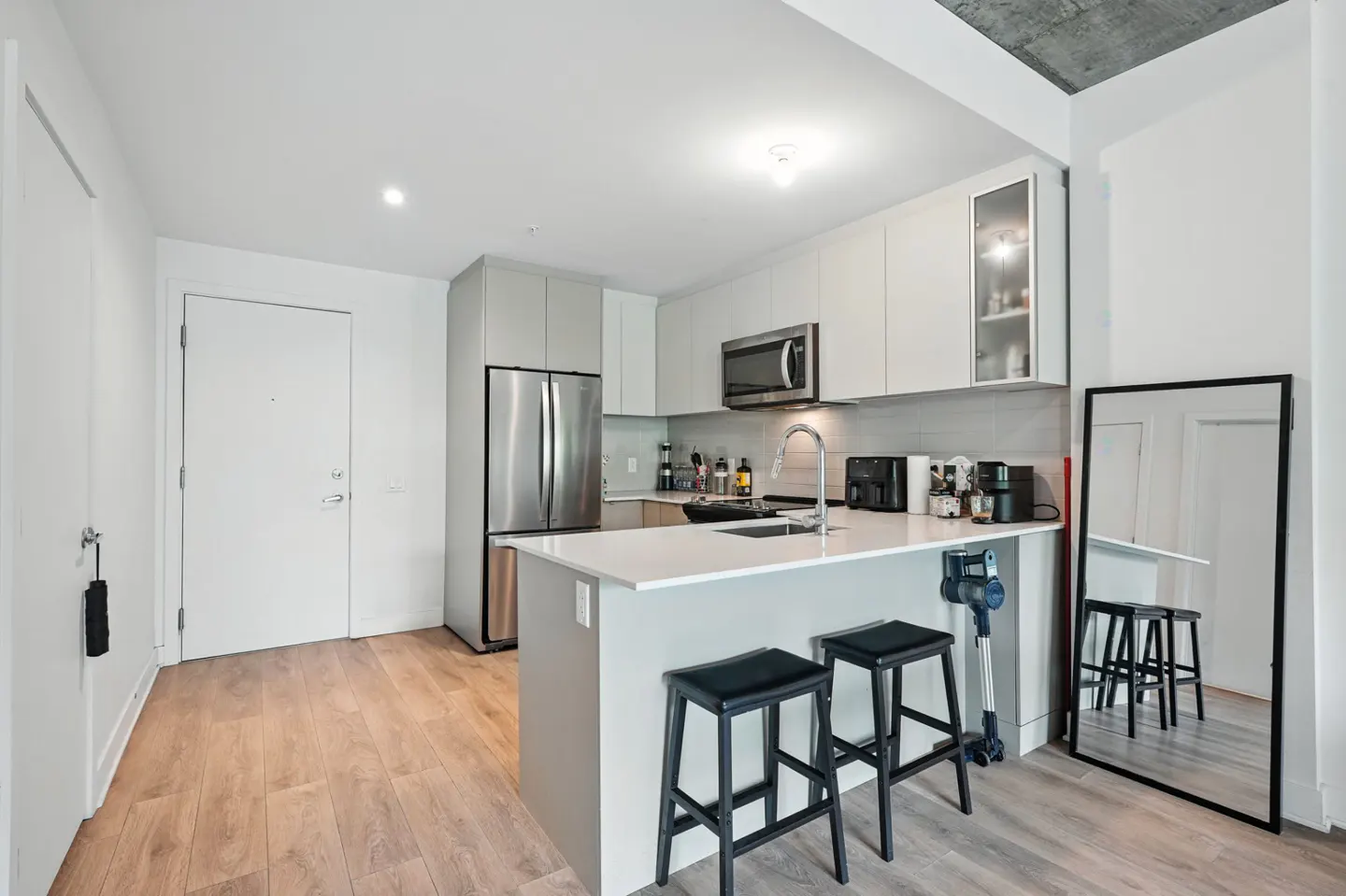 Modern kitchen with white cabinets, stainless steel appliances, and a white countertop island with two black stools. A black framed mirror reflects the kitchen.
