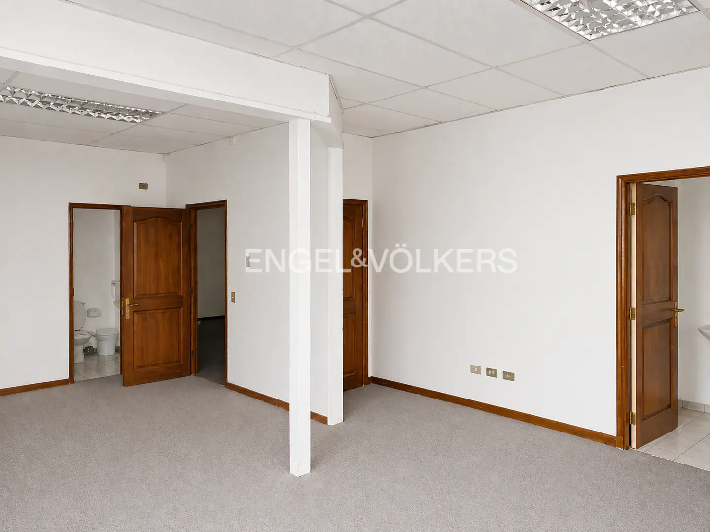 An empty room with gray carpet, white walls, and brown doors. The Engel & Völkers logo is on the wall.