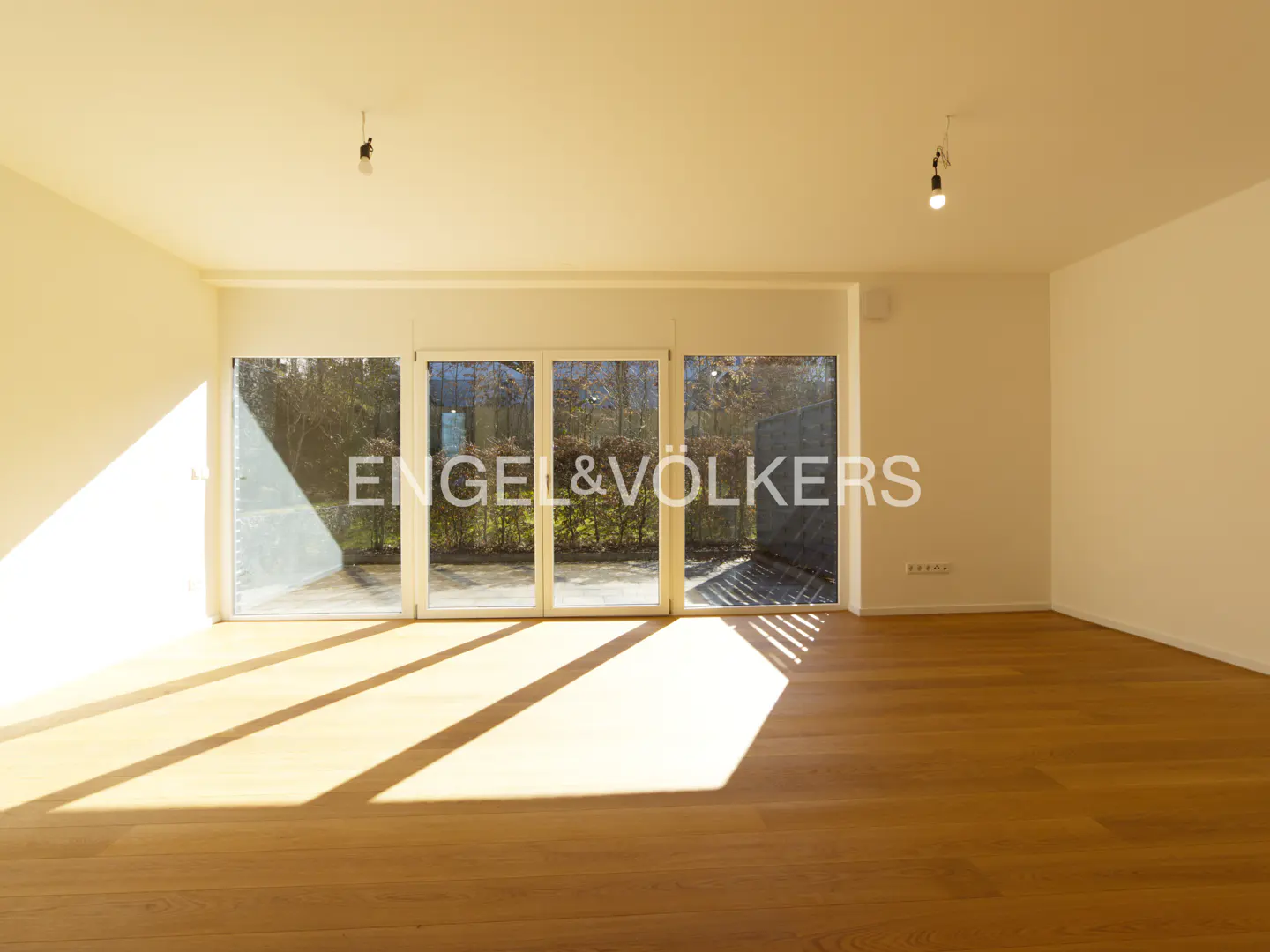 Bright, empty room with hardwood floors and white walls. Sunlight streams in through glass doors to a patio. Engel & Völkers logo visible.