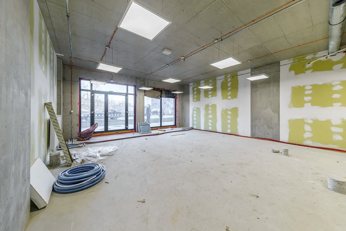 Unfinished commercial space with concrete ceiling, exposed pipes, drywall patches, and blue conduit coils on the floor.