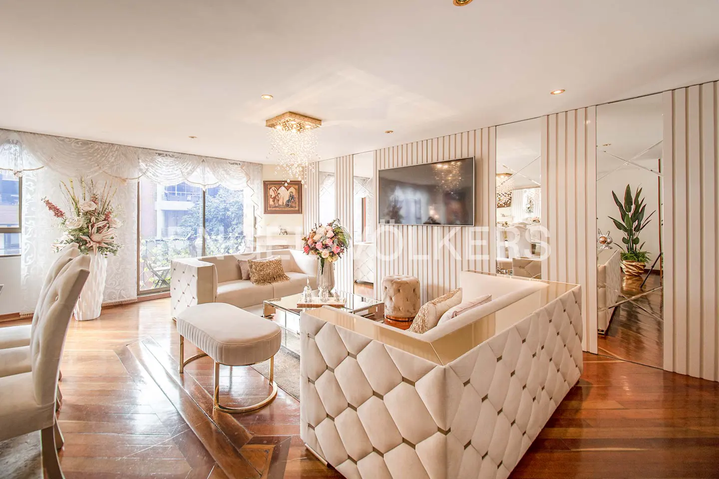 A bright living room with beige sofas, a glass coffee table, and hardwood floors. A TV hangs on a striped wall with mirrors.