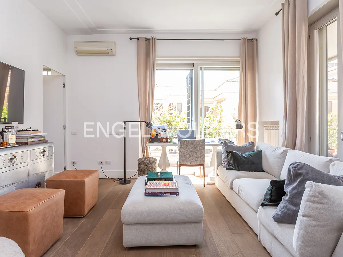 Bright living room with a white sofa, grey ottoman, two tan leather poufs, and a desk by a window with beige curtains.
