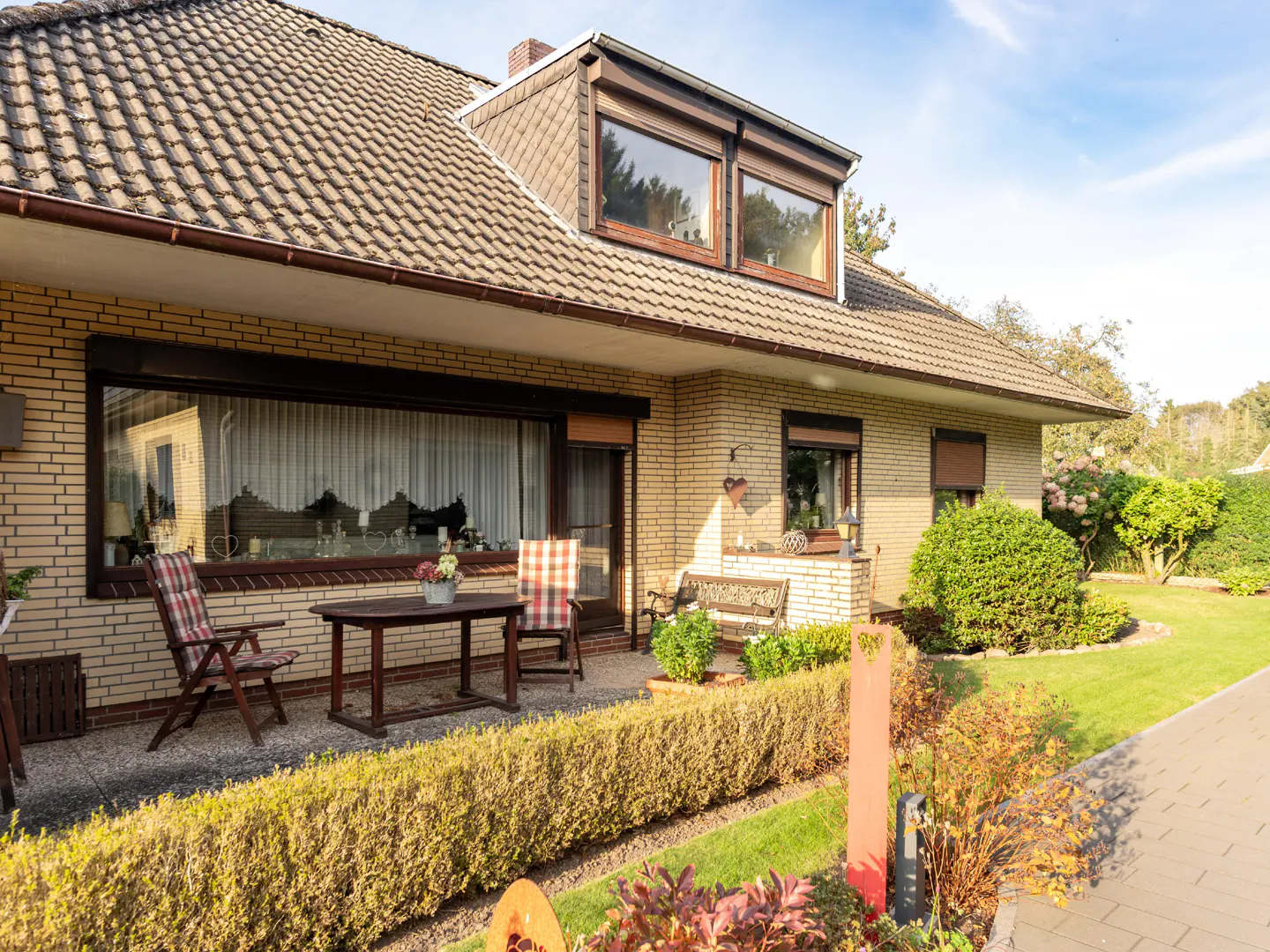 A single-story brick house with a brown tiled roof, a patio with chairs and a table, and a green lawn.
