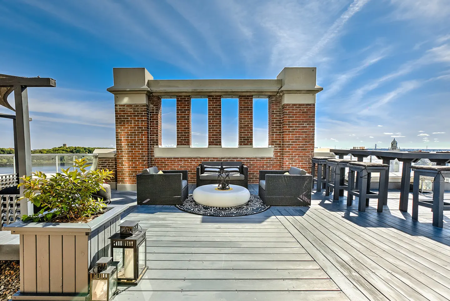 Rooftop patio with gray wood planks, brick wall with three window openings, black wicker furniture, and a white round table.