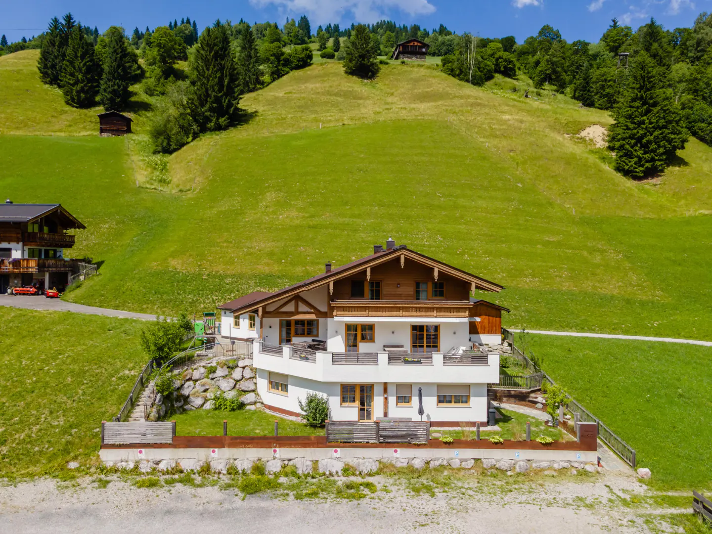 A two-story white house with a brown roof sits on a green hillside with trees and other houses in the background.