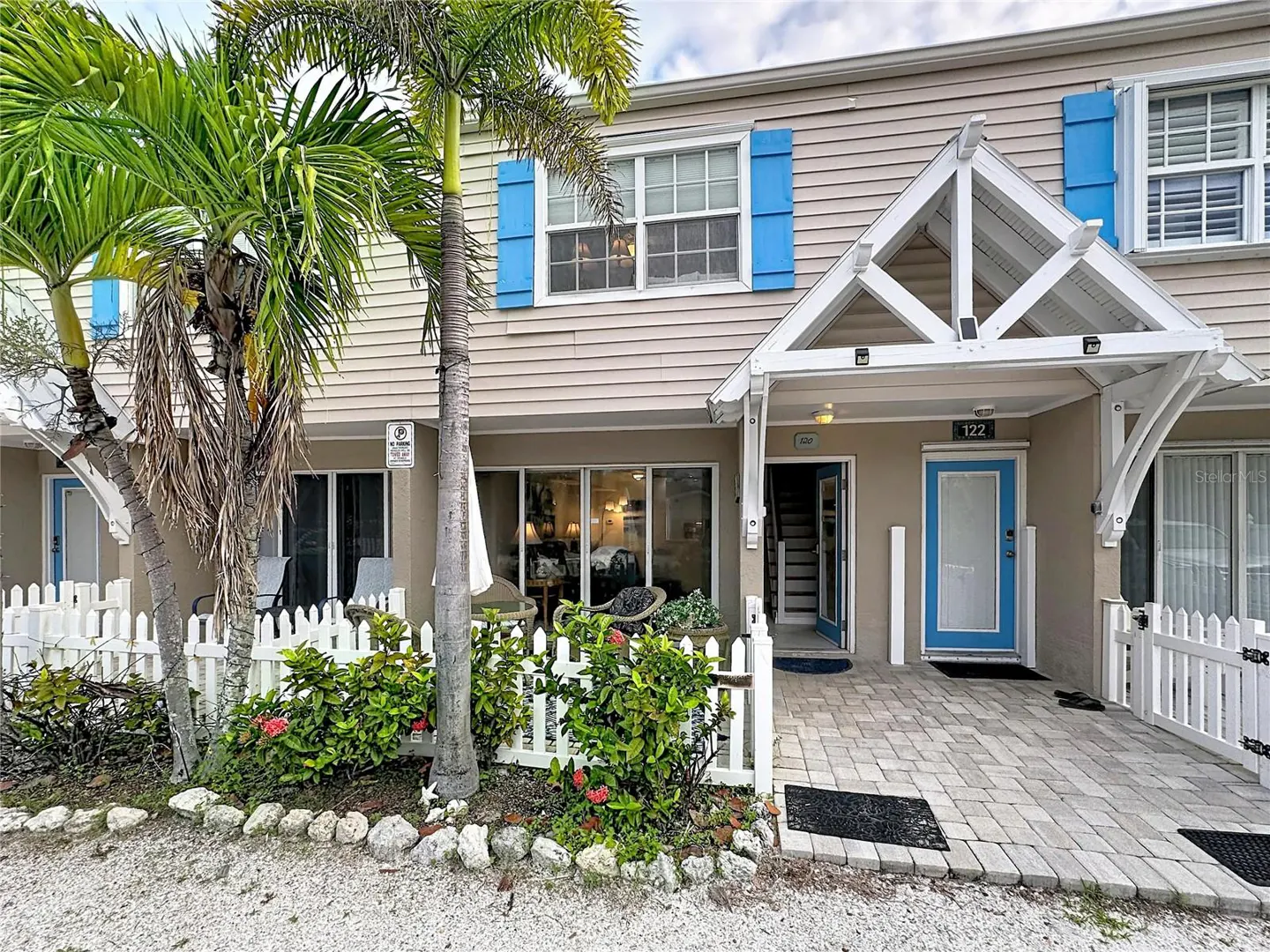 Tan townhouse with blue shutters, a white front porch, and a palm tree in front of a white picket fence.