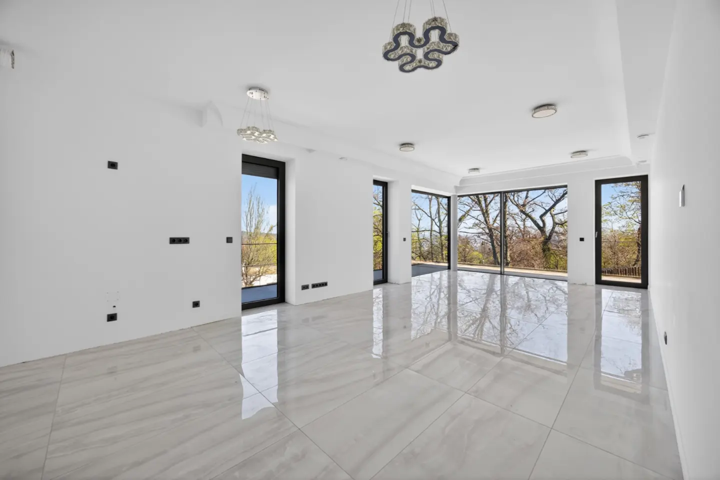 Bright, empty room with white walls, marble floors, and black-framed windows showing trees. Modern chandeliers hang from the white ceiling.