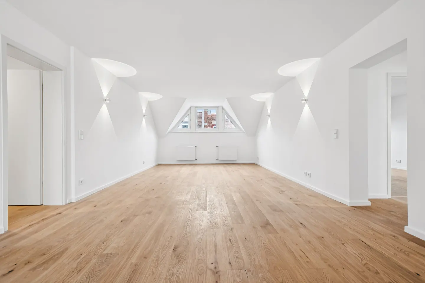 Empty attic room with white walls, light wood floors, sloped ceilings, wall sconces, and a central window.