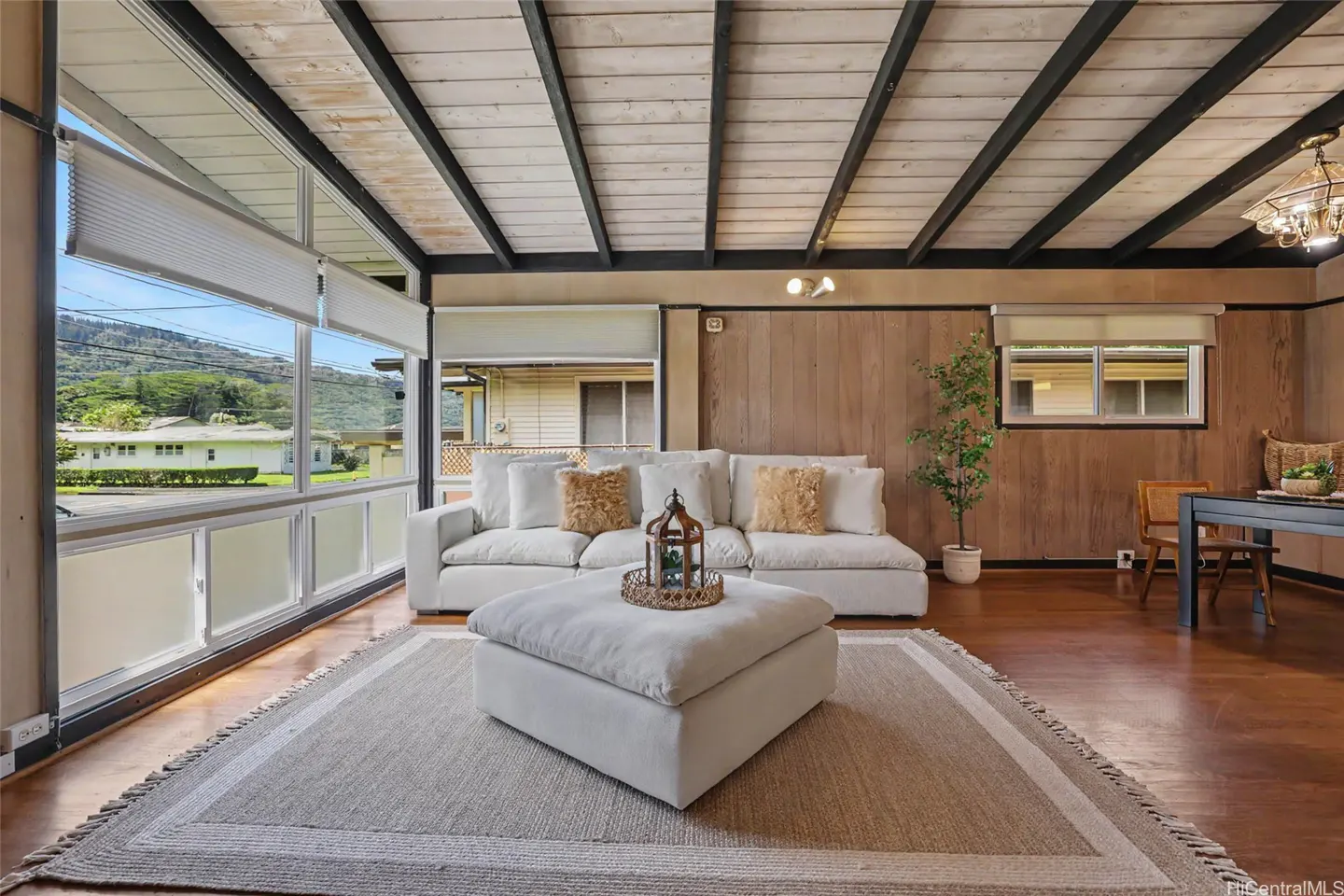 Living room with white sofa, ottoman, and wood-paneled walls. Large windows offer a view of a green landscape.