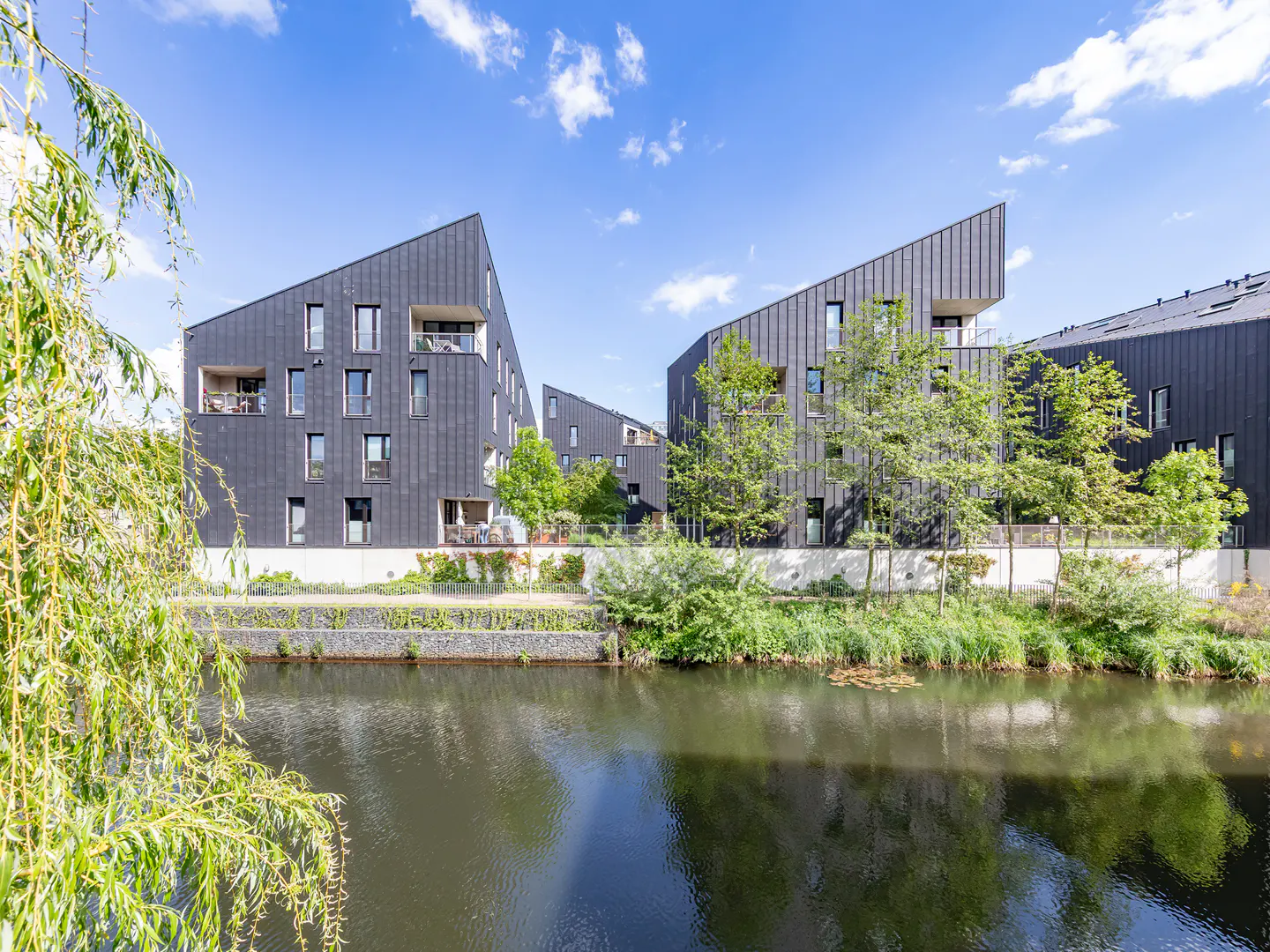 Modern black apartments with angular roofs line a canal under a blue sky with white clouds. Green trees and foliage surround the buildings.