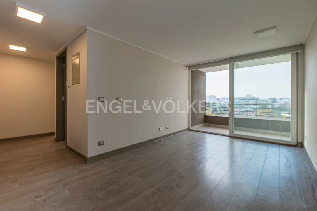 An empty room with gray wood floors, white walls, and a large window with a balcony overlooking a city. The Engel & Völkers logo is on the wall.