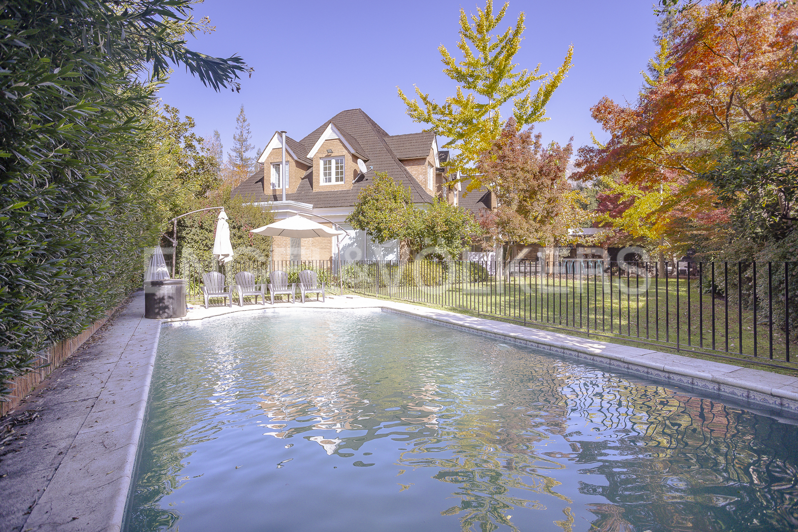 A backyard pool reflects a brick house with a brown roof and autumn trees under a blue sky. [AI-generated text]