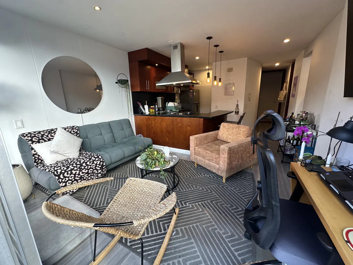 Modern open-plan living room with a grey sofa, tan armchair, woven rocking chair, and a kitchen with dark wood cabinets.