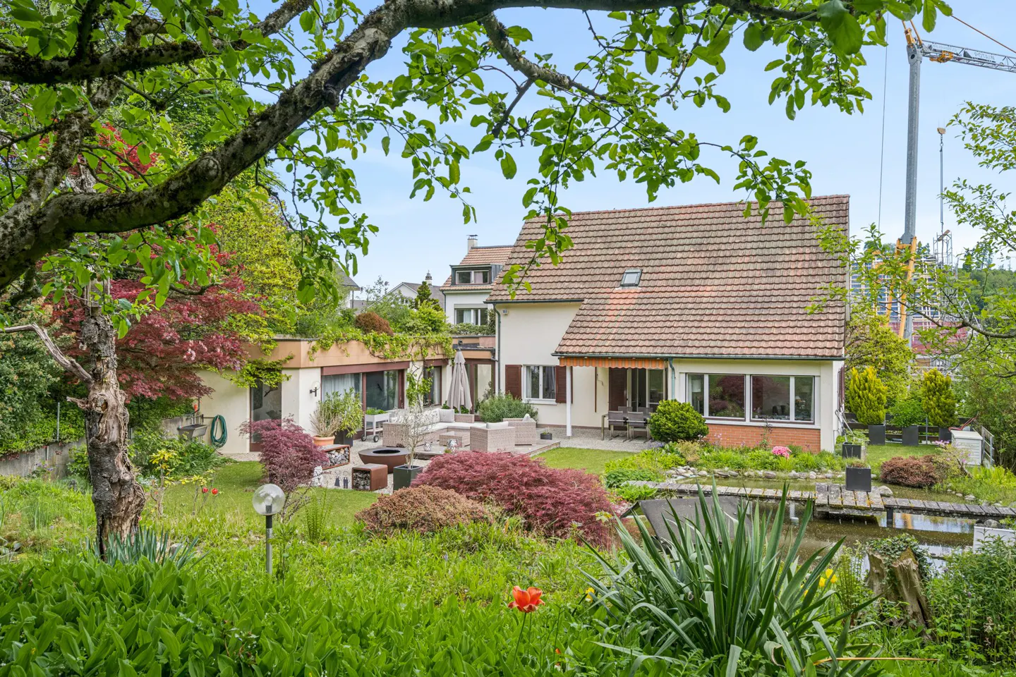 Exterior view of a house with a red tile roof, a lush green garden, and a pond. Outdoor furniture is visible on the patio.