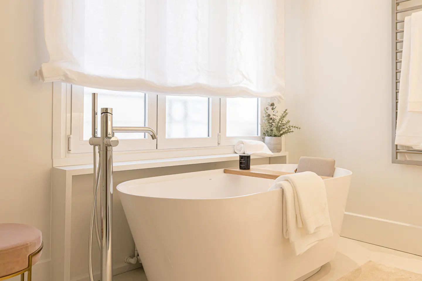 Modern white freestanding bathtub with chrome faucet, wooden tray, and white towels in a bright, minimalist bathroom.