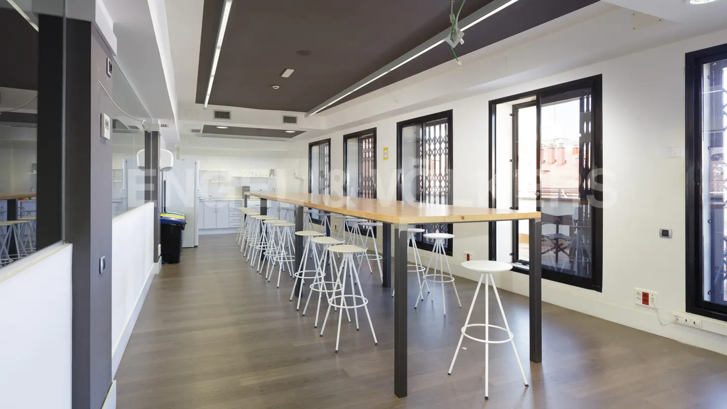 Bright office space with a long wooden table and white stools. Windows with black frames line the right wall. A kitchen area is visible in the background.