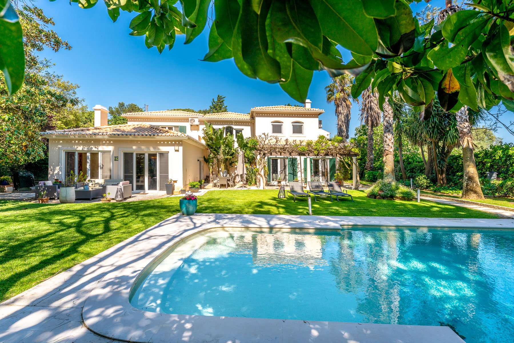 A sunny backyard with a swimming pool, green lawn, patio furniture, and a two-story cream-colored villa under a blue sky. [AI-generated text]