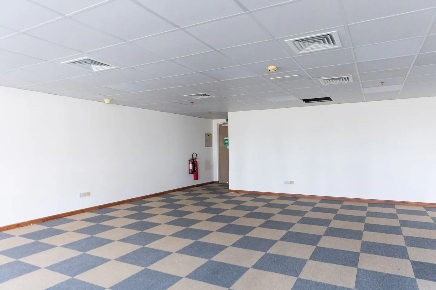 Empty office space with white walls, checkered blue and beige carpet, and a white tile ceiling. A red fire extinguisher hangs near an exit door.