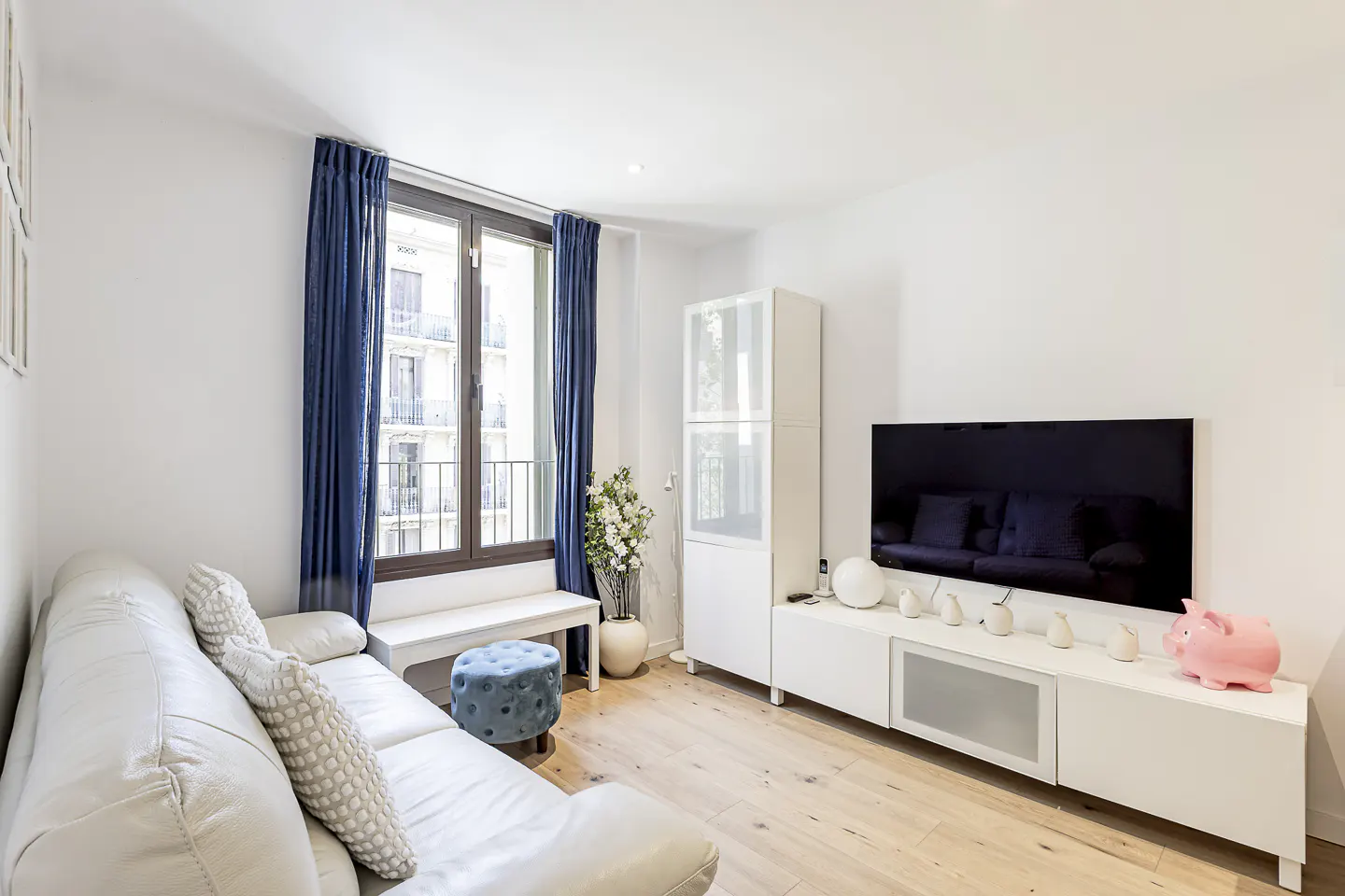 Bright living room with white walls, wood floors, and blue curtains. A white sofa faces a TV, with a pink piggy bank on the console.