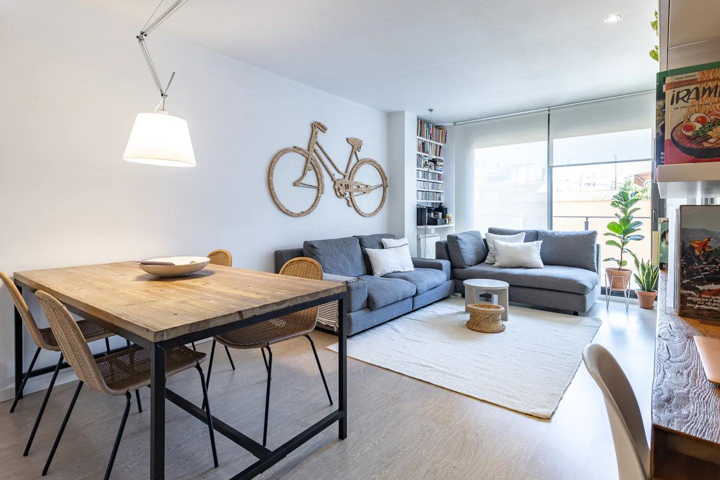 Bright living room with a wooden table, wicker chairs, and a gray sofa. A decorative bicycle hangs on the white wall.