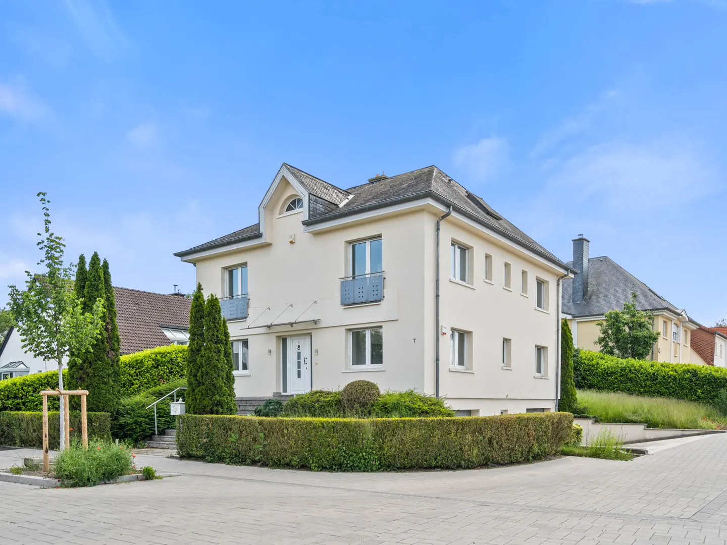 Two-story cream house with a gray roof, white trim, and manicured green hedges under a blue sky.