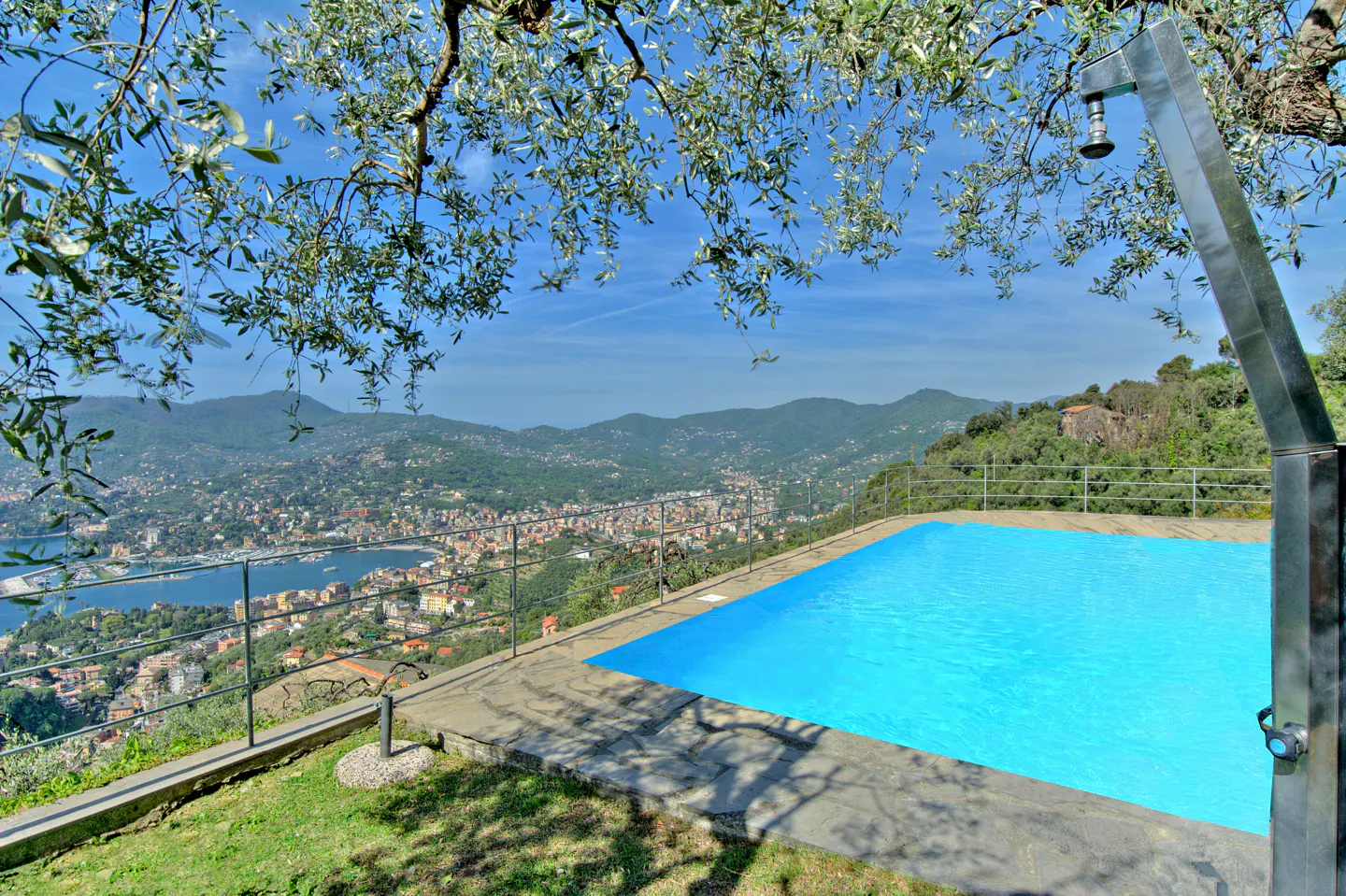 Outdoor pool with blue water overlooks a city, mountains, and the sea. A metal shower stands next to the pool.