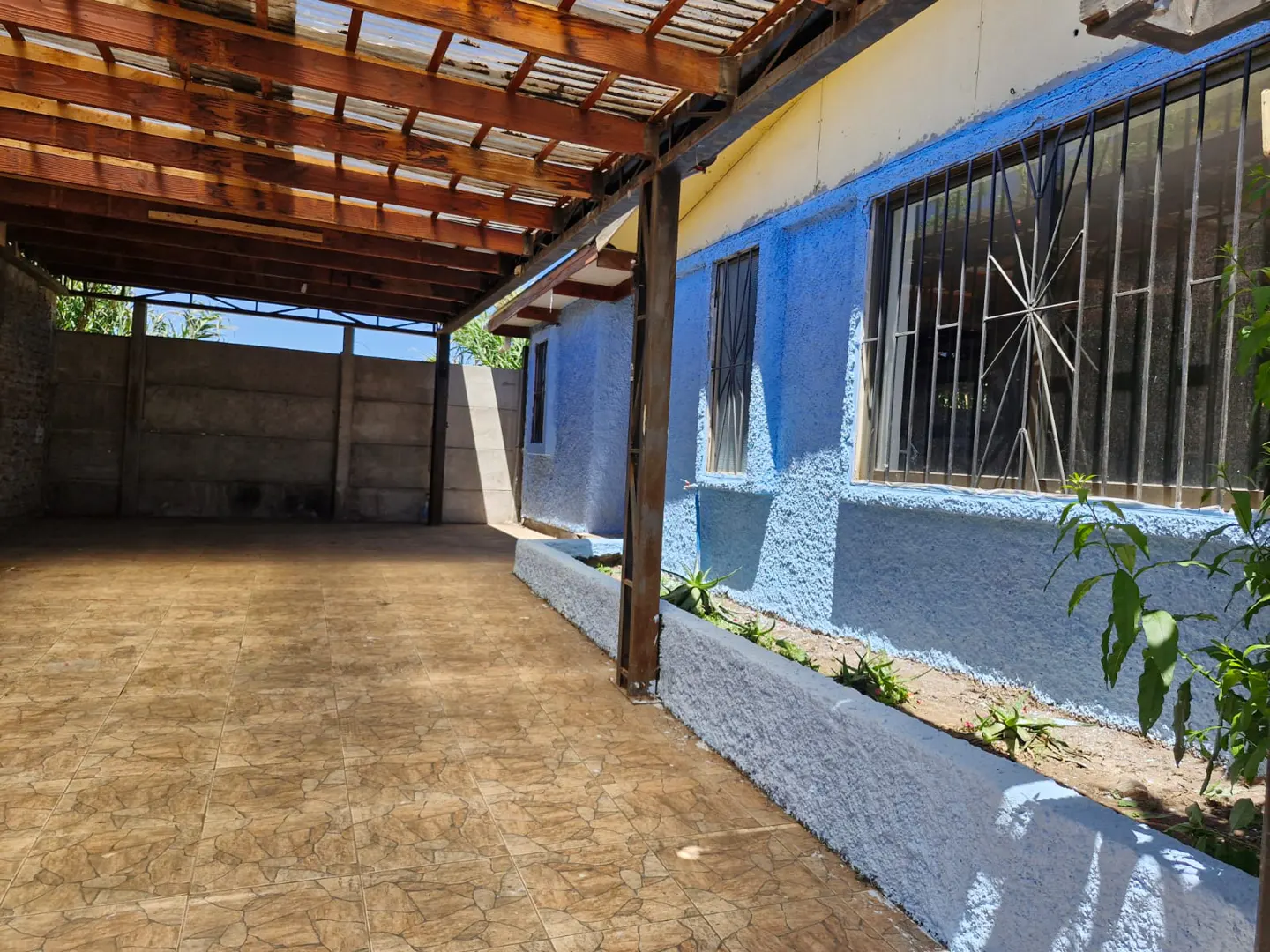 Covered patio with tiled floor, blue textured wall, and a raised garden bed next to a house with barred windows.