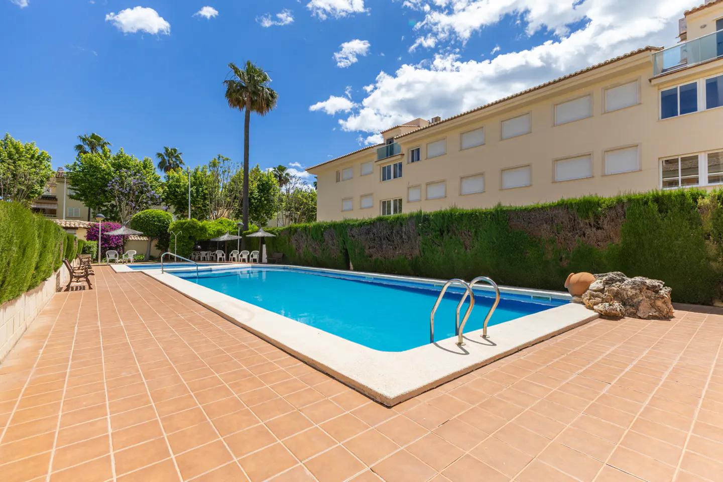 Outdoor pool with blue water, surrounded by a tiled patio, green hedges, and a beige building under a blue sky.