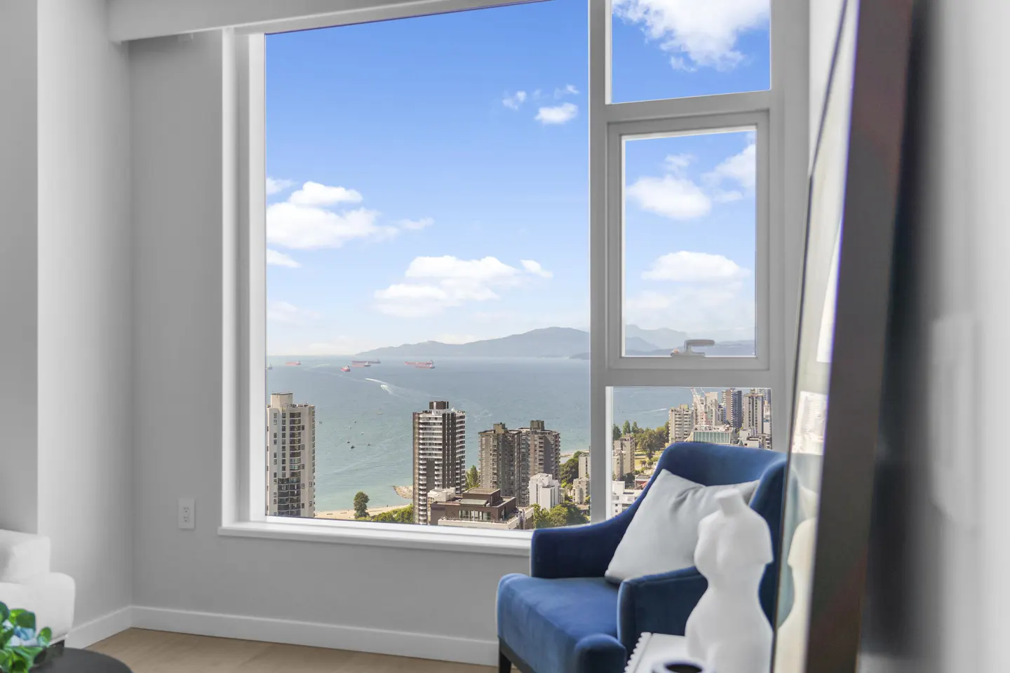 Bright room with a blue chair facing a large window. Ocean view with ships, mountains, and city buildings visible.