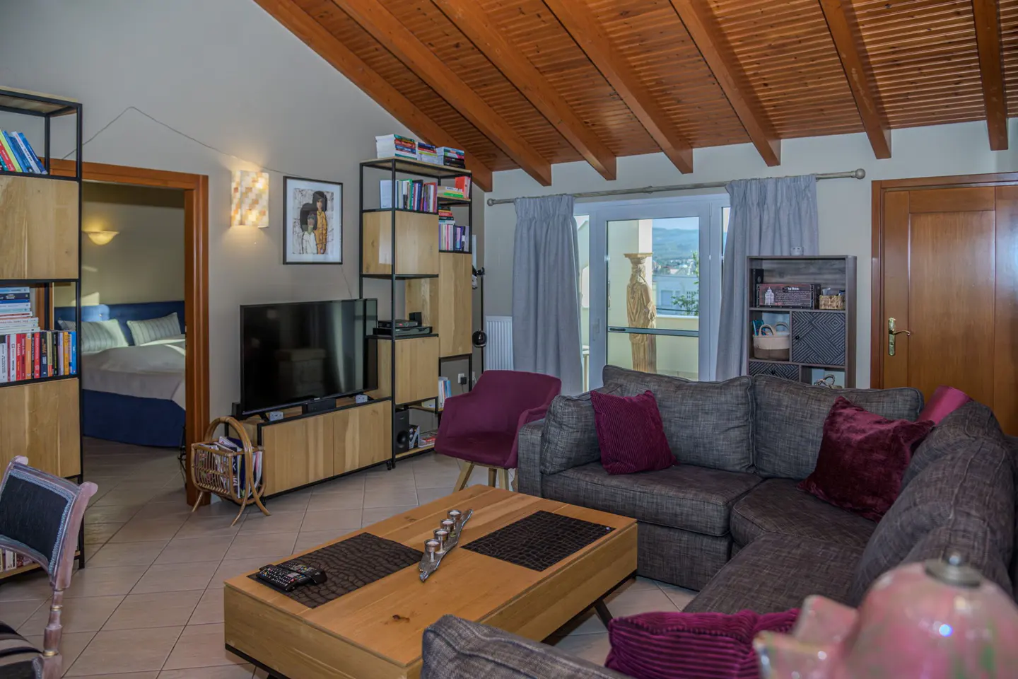 A cozy living room with a wood-beamed ceiling, gray sectional sofa with purple pillows, and a wooden coffee table. A TV and bookshelves are against the wall.