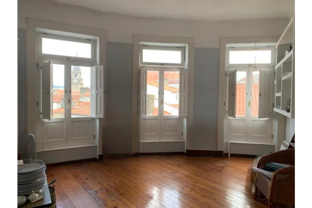 A bright room with wood floors and three white-framed windows with open shutters, offering a city view.