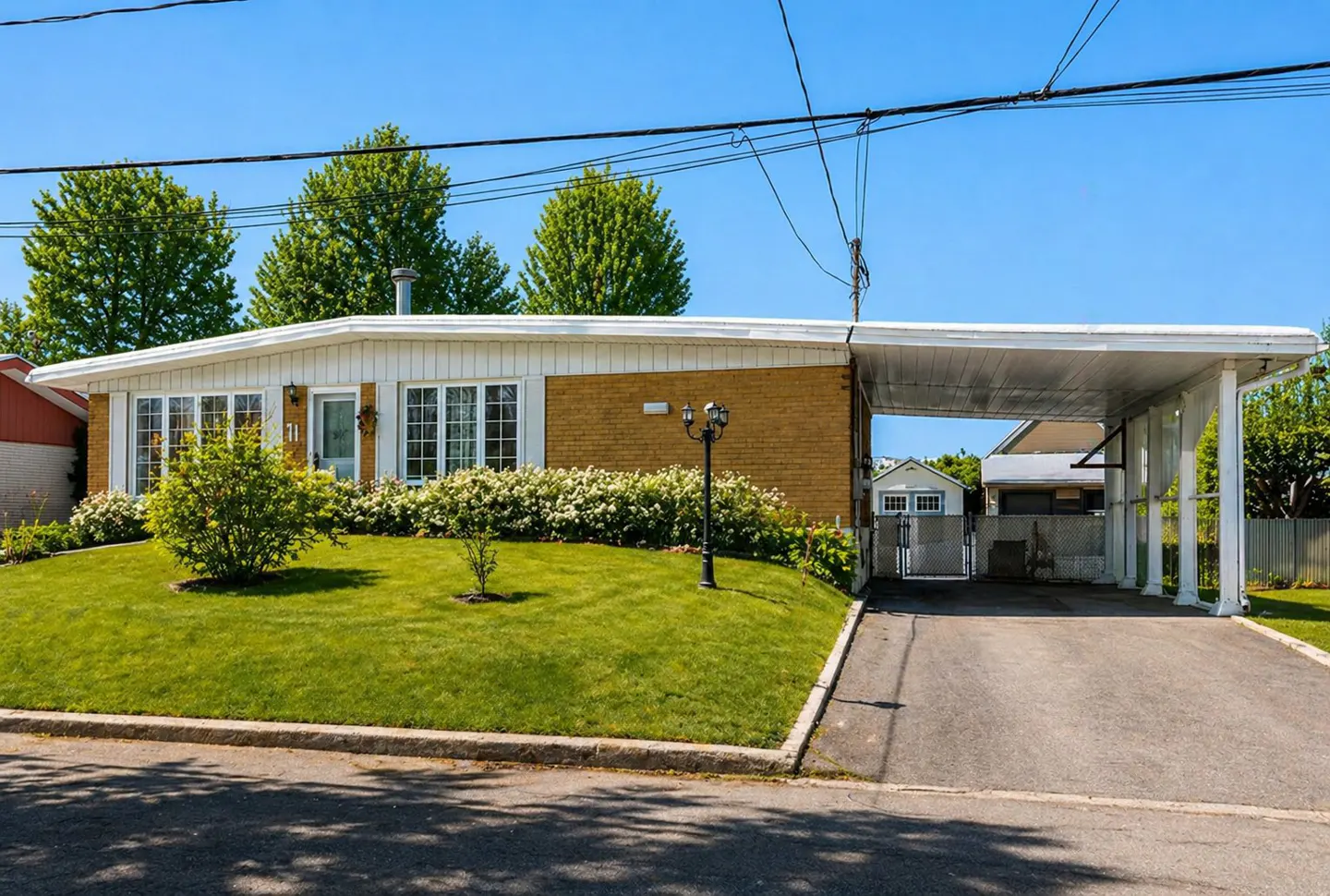 Single-story yellow brick house with a white carport, green lawn, and flowering bushes under a clear blue sky.