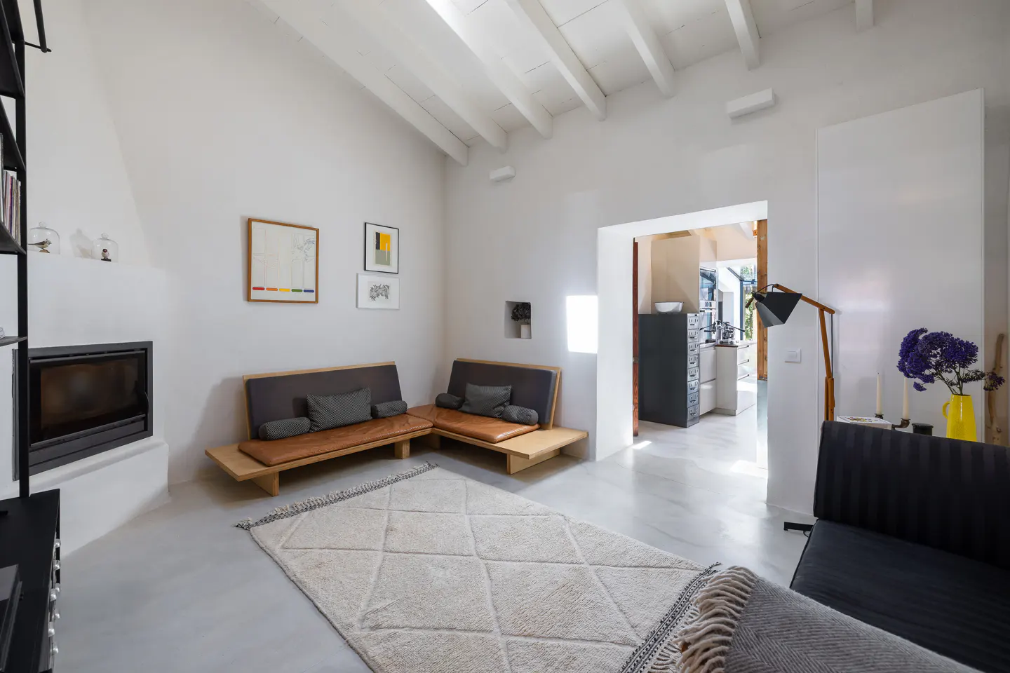 Bright living room with white walls, wooden sofas, and a diamond-patterned rug. A fireplace and artwork adorn the walls. A doorway leads to a kitchen.