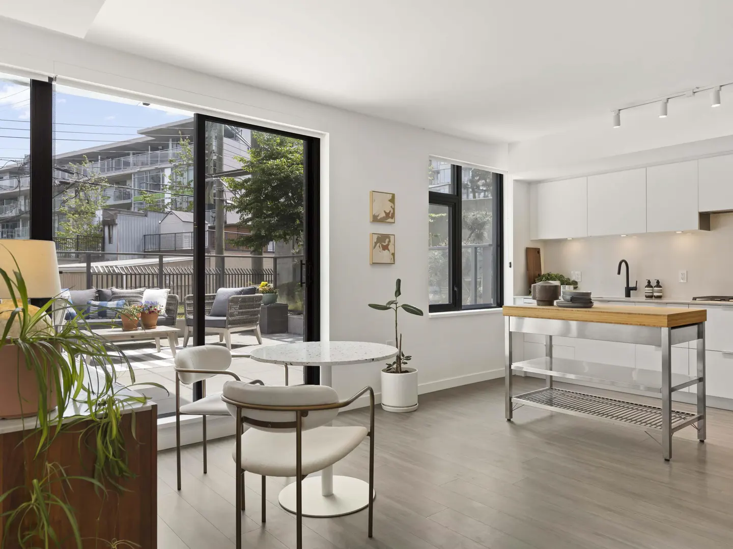 Bright, modern apartment interior with white walls, wood floors, and a kitchen island. A patio with furniture is visible through a large sliding glass door.