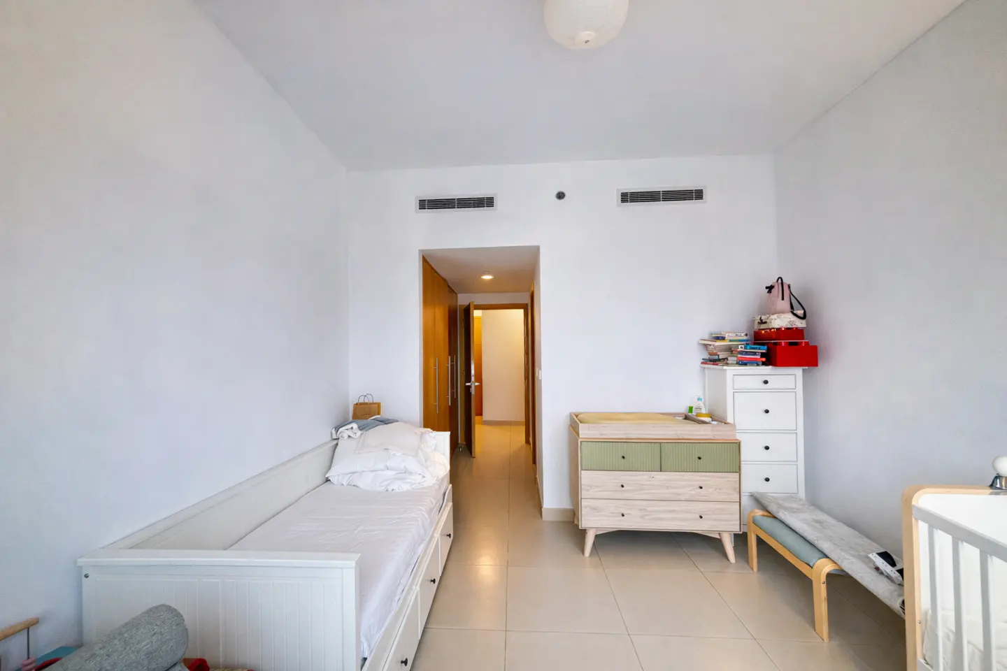 Bright, white bedroom with a daybed, dresser, and open doorway. A white dresser with books and a red box sits near a changing table.