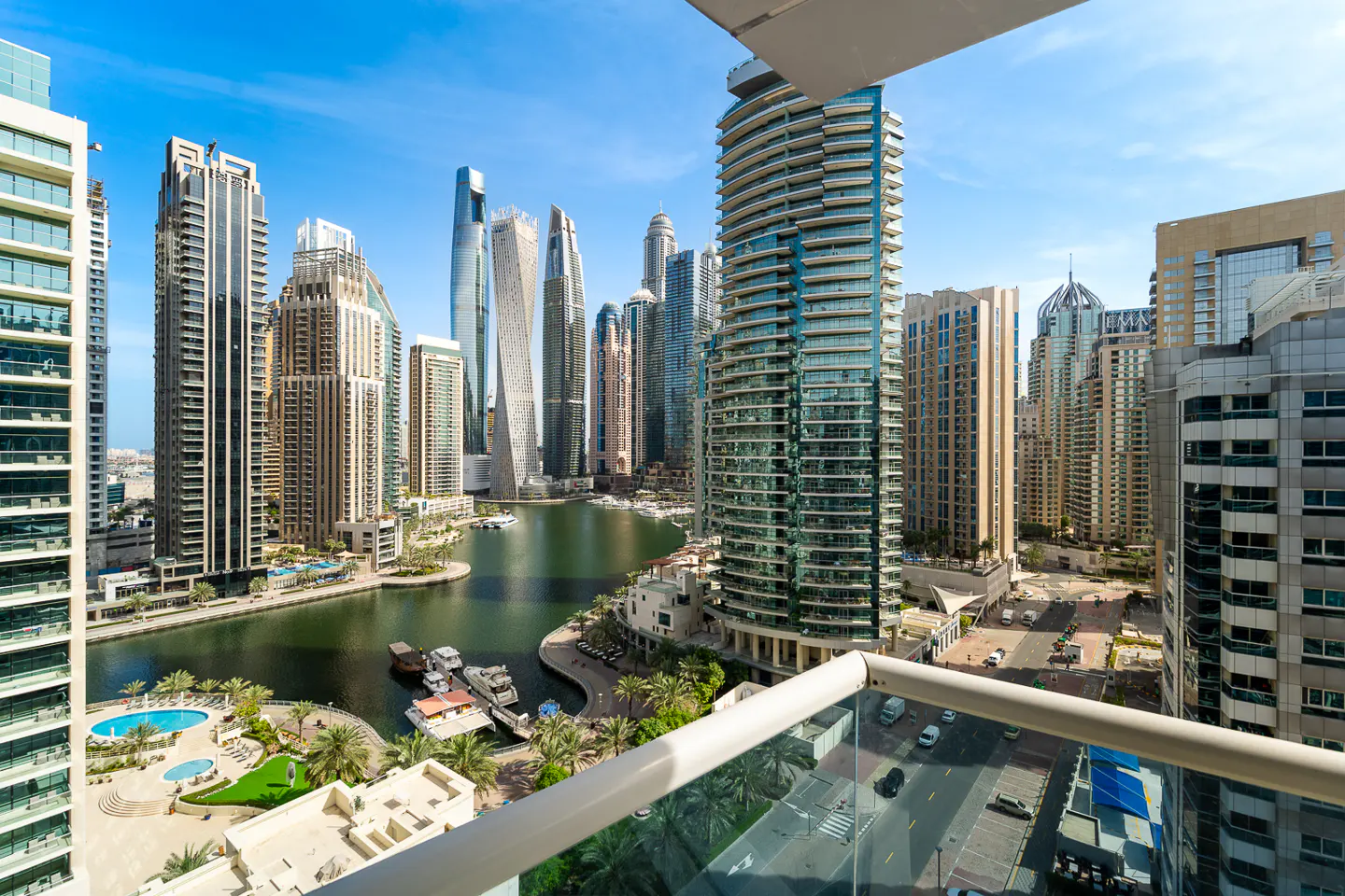 View from a balcony overlooking Dubai Marina, with skyscrapers, water, boats, and a street with cars visible.