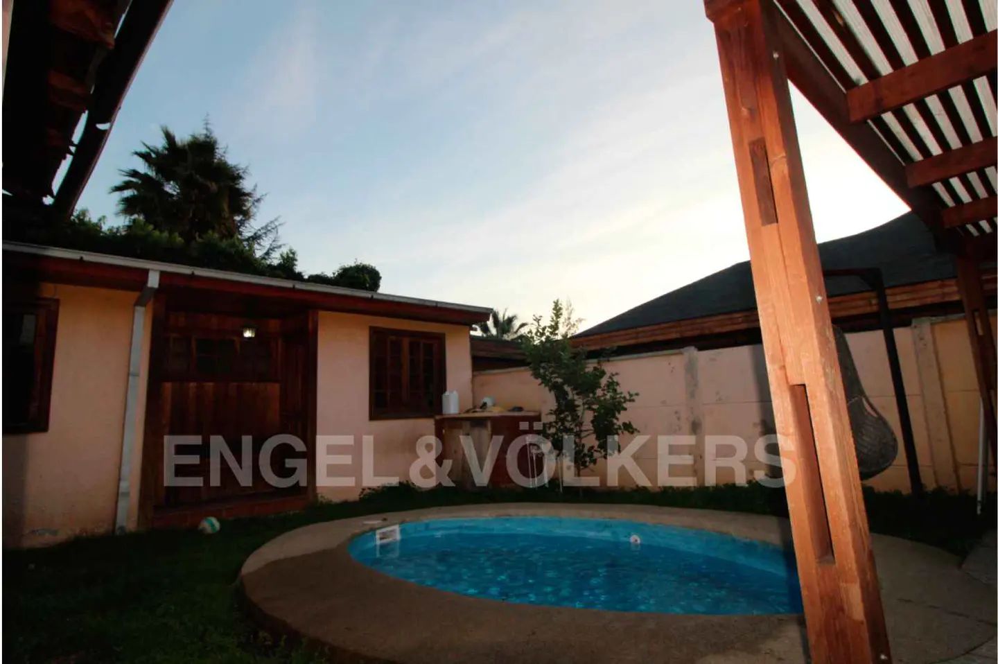 Backyard view of a small, round, blue pool with a tan house, wooden pergola, and green grass.
