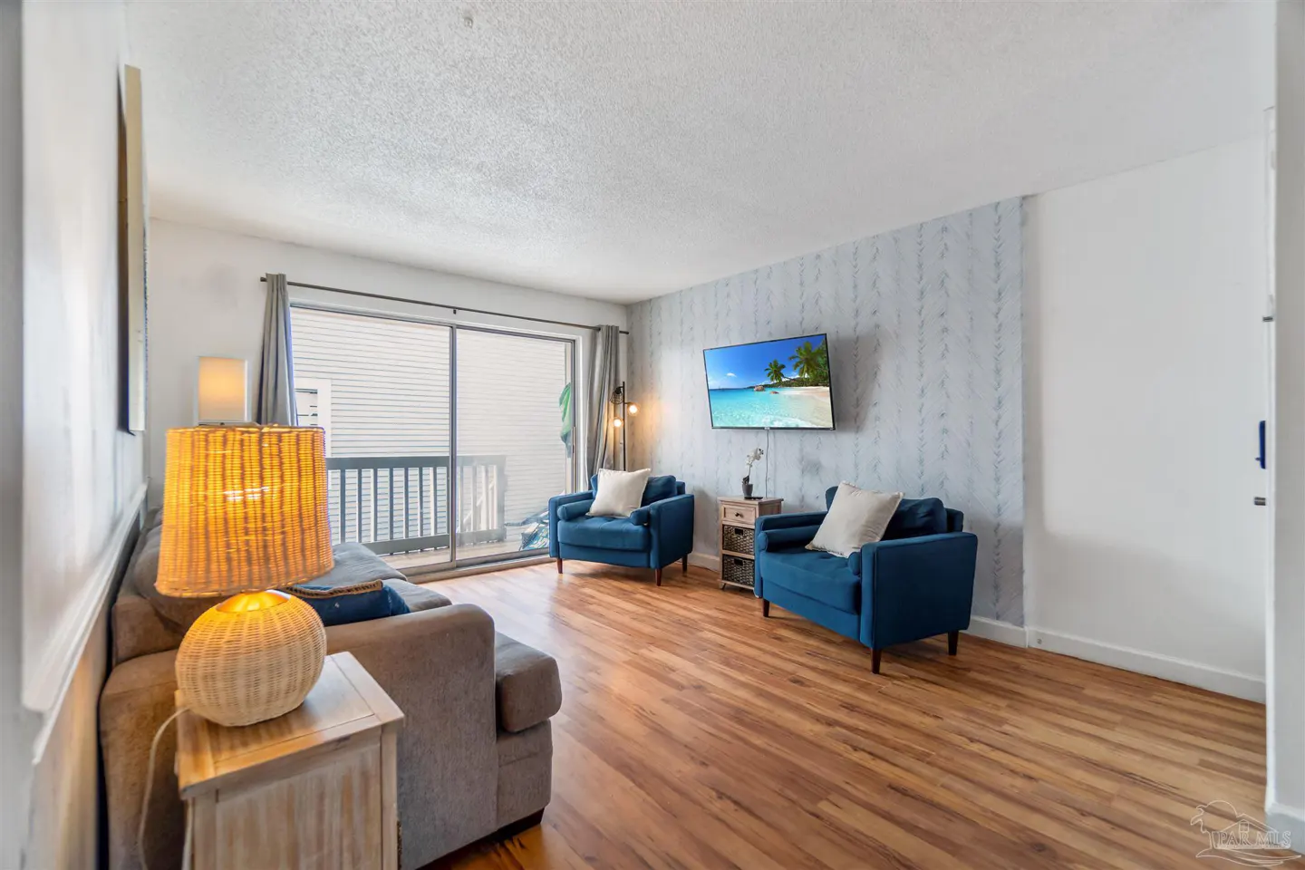 Living room with wood floors, a gray sofa, two blue armchairs, and a TV showing a beach scene. Sliding glass doors lead to a balcony.