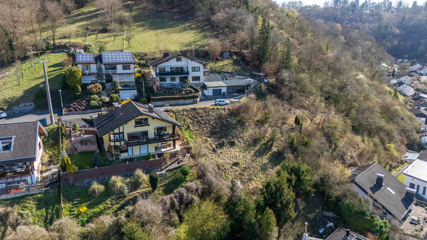 Aerial view of hillside homes, one with yellow siding and a balcony, surrounded by trees and greenery.