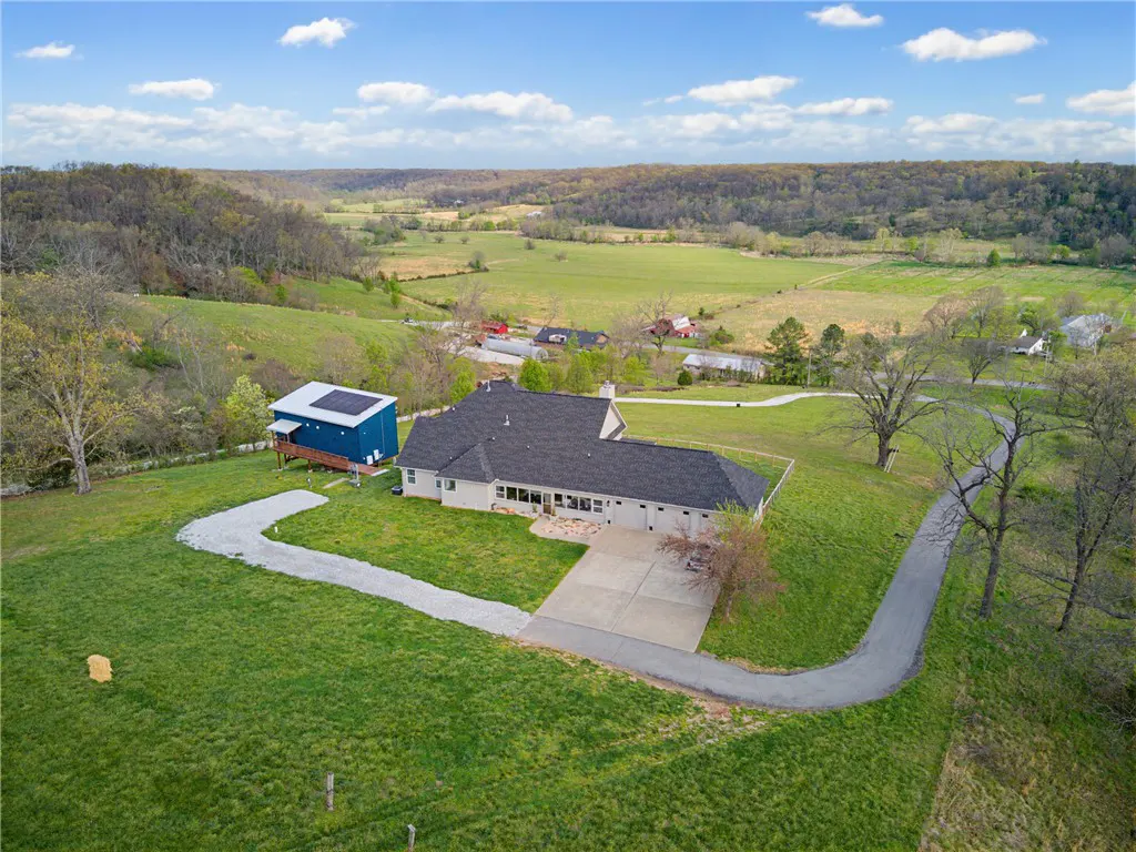 Aerial view of a ranch-style house with a dark roof, a blue outbuilding with solar panels, and a long driveway in a green, hilly landscape.