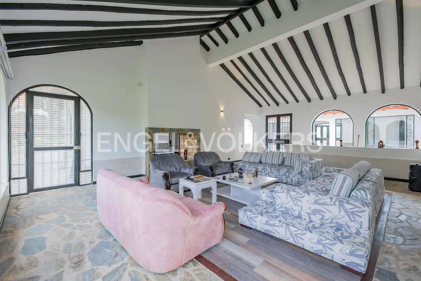 Bright living room with stone floors, white walls, and dark wood beams. Sofas and chairs surround a white coffee table. Arched windows and doors let in natural light.