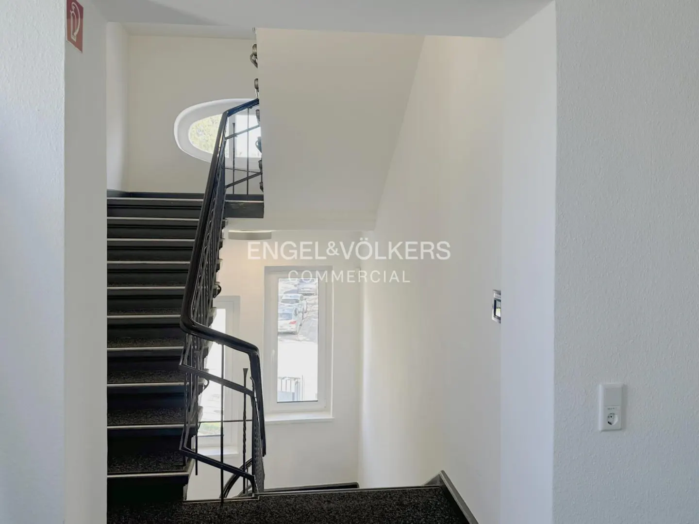 A bright stairwell with dark steps, a black metal railing, white walls, and an oval window overlooking a parking lot.