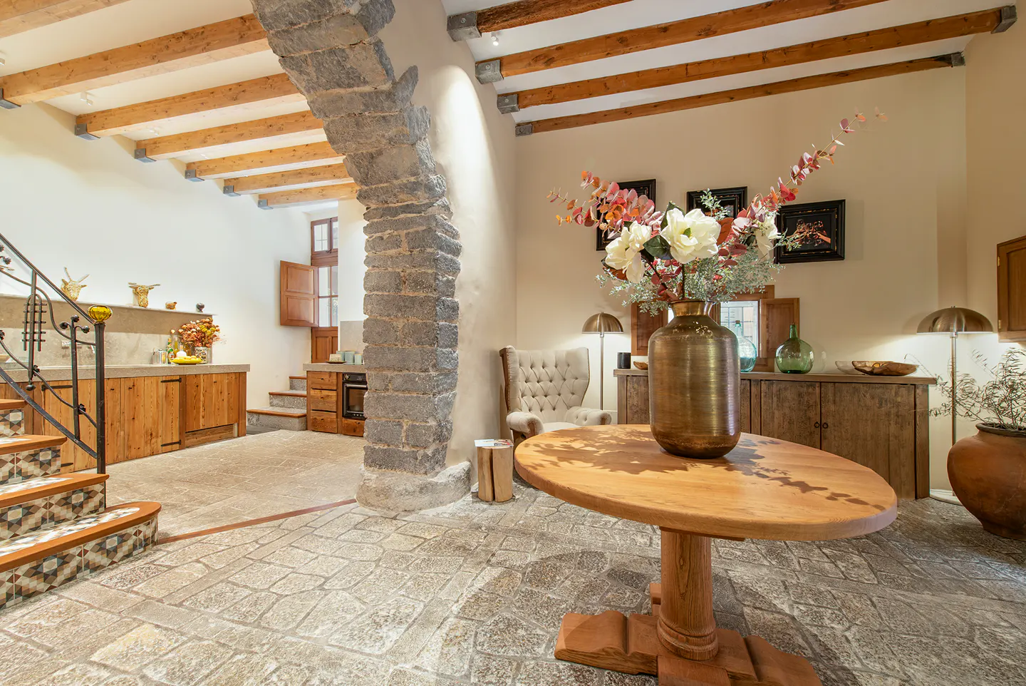 Interior view of a rustic home with stone floors, wooden beams, and a stone archway. A round wooden table with flowers sits in the foreground.