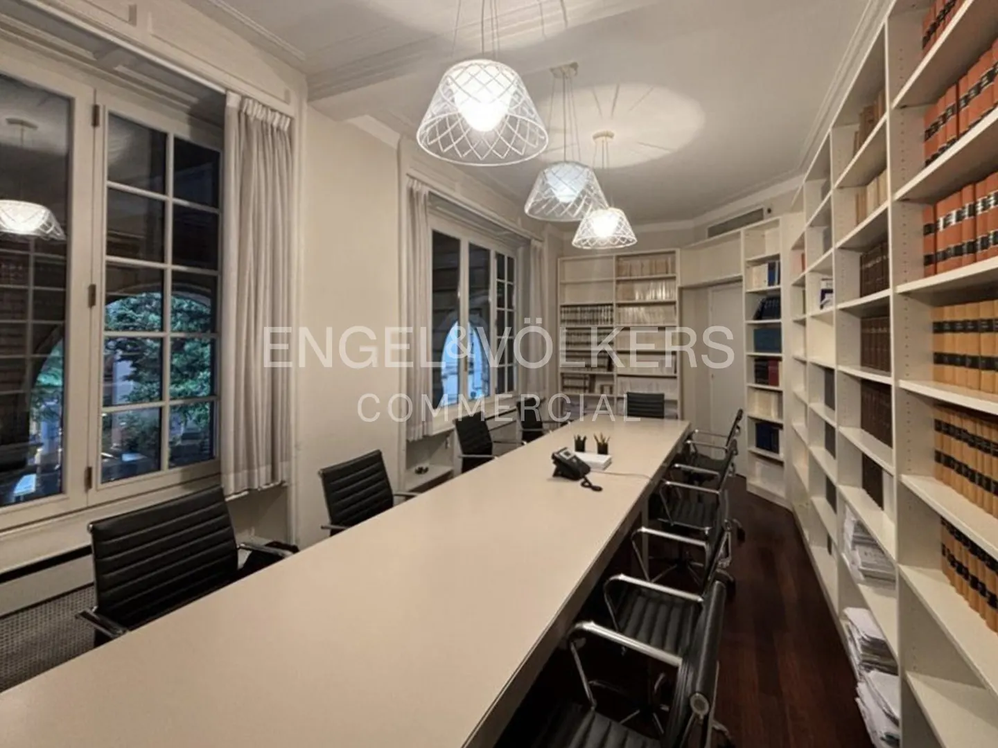 A bright conference room with a long table, black chairs, and white bookshelves filled with books. Three modern pendant lights hang above.