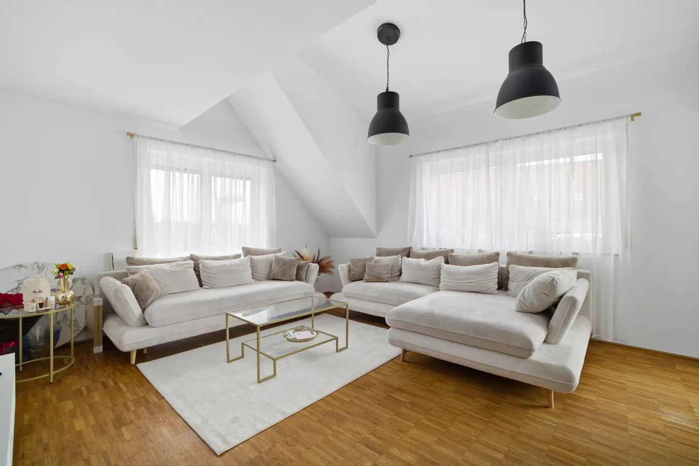 Bright living room with two beige sofas, a glass coffee table, and wood floors. White walls and sheer curtains add to the airy feel.