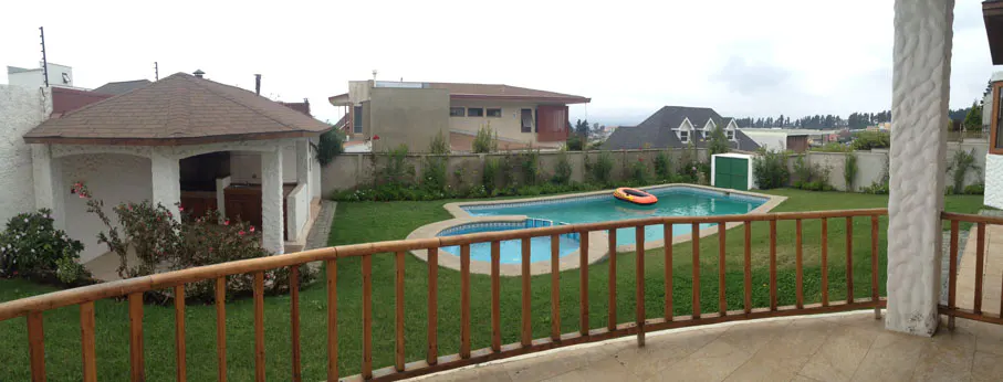 Backyard view from a patio with a wooden railing, showing a pool with an orange raft, a gazebo, and green grass.